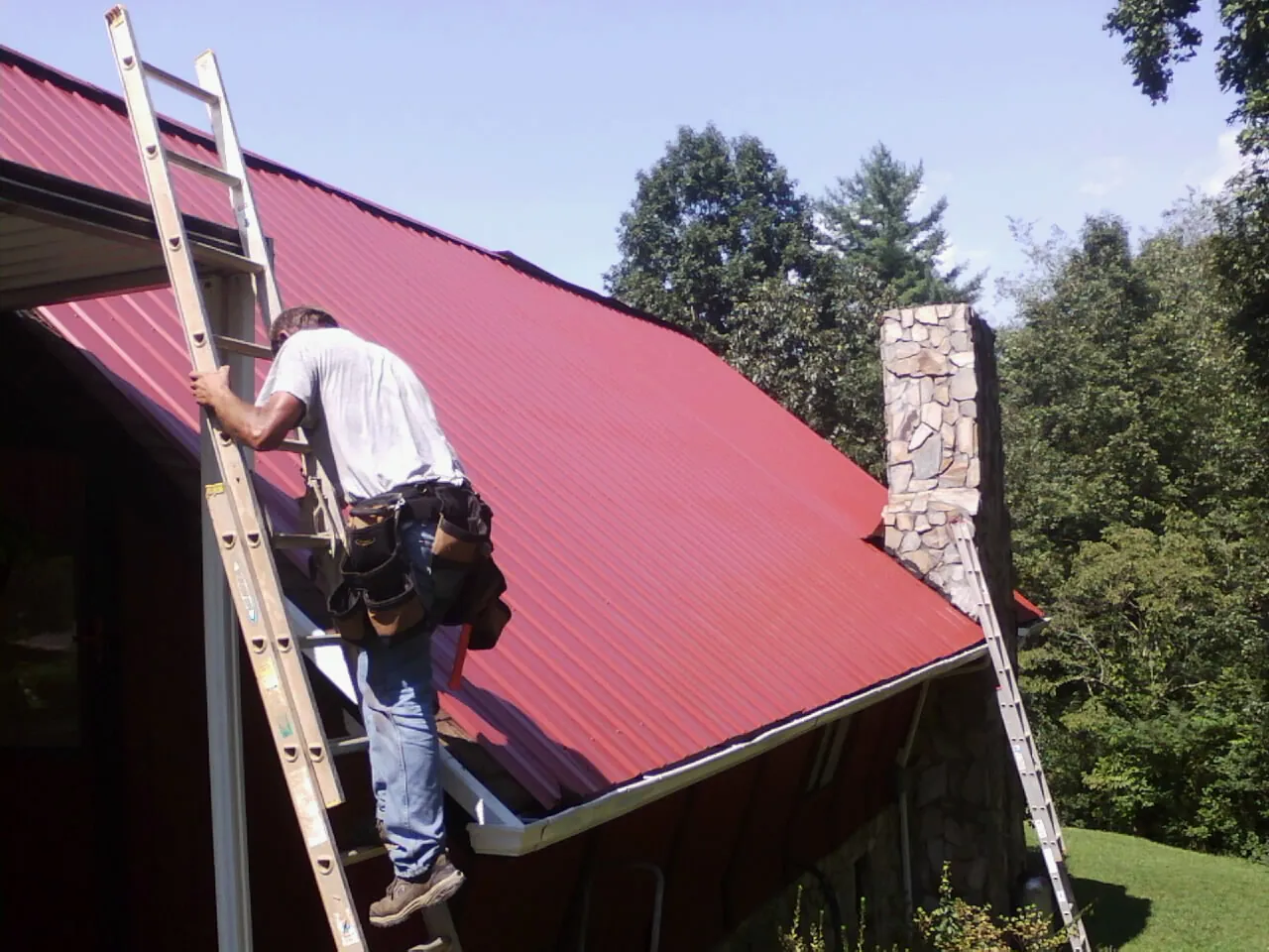 Roofer on a ladder beside a red metal roof and stone chimney — Soffit & Fascia Repair by Roofing Brothers in San Juan Bautista